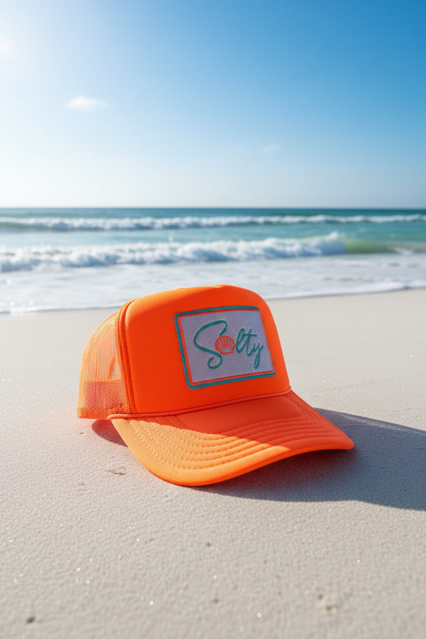 Bright orange trucker hat with a custom embroidered patch on the front featuring a white background, teal and silver glitter border, and teal and orange lettering.