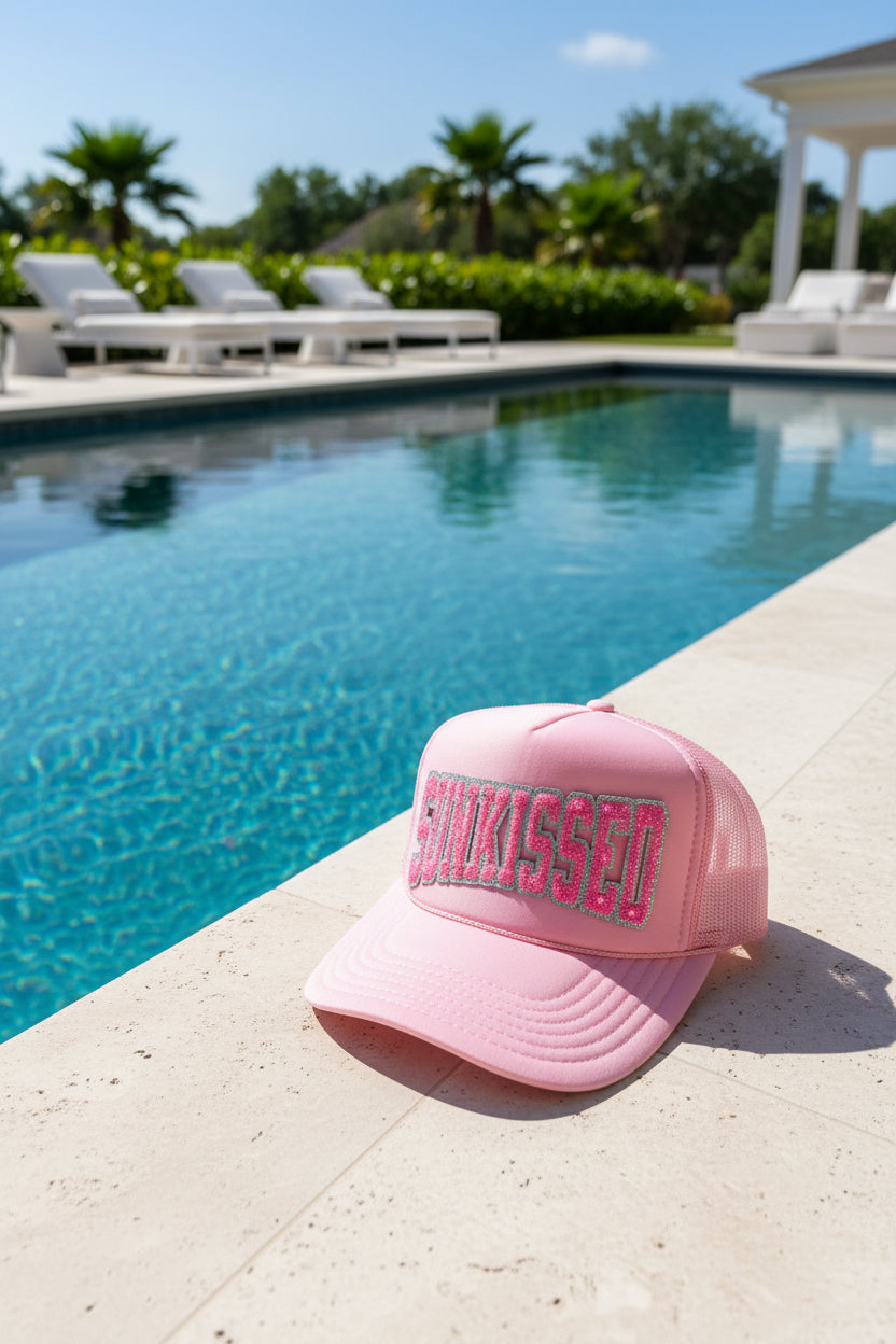 A pink trucker hat with mesh back panels and a curved brim, featuring the word 'Sunkissed' in glittery pink letters outlined in silver on the front panel, resting on a light-colored surface.