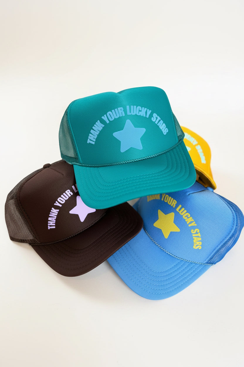 Three trucker hats stacked together on a gray textured surface. The top hat is teal with a light blue star and white text reading 'THANK YOUR LUCKY STARS.