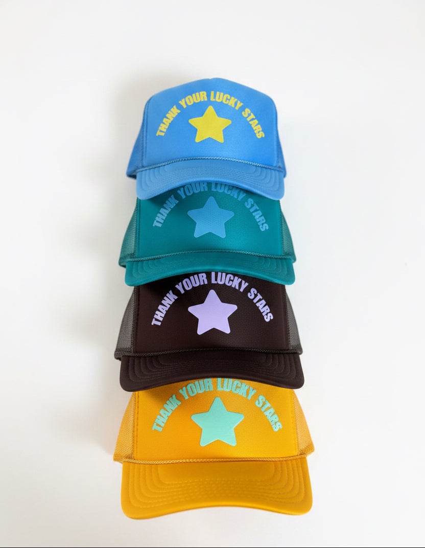 Five trucker-style caps stacked vertically on a gray carpeted floor. Each cap features a different bright color (blue, teal, brown, yellow, and orange) with a star graphic and the text 'THANK YOUR LUCKY STARS' printed on the front.