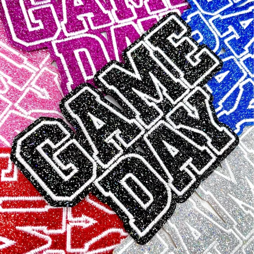 A close-up of a glittery 'GAME DAY' patch or decal with black glitter lettering outlined in white, layered over a colorful background with pink, blue, red, and silver glitter text.