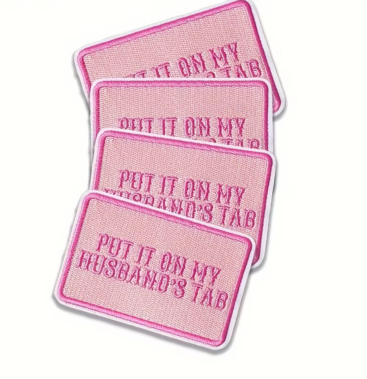 Four pink rectangular embroidered patches with rounded corners, stacked diagonally on a white background. Each patch reads 'PUT IT ON MY HUSBAND'S TAB' in darker pink embroidery with a white border.