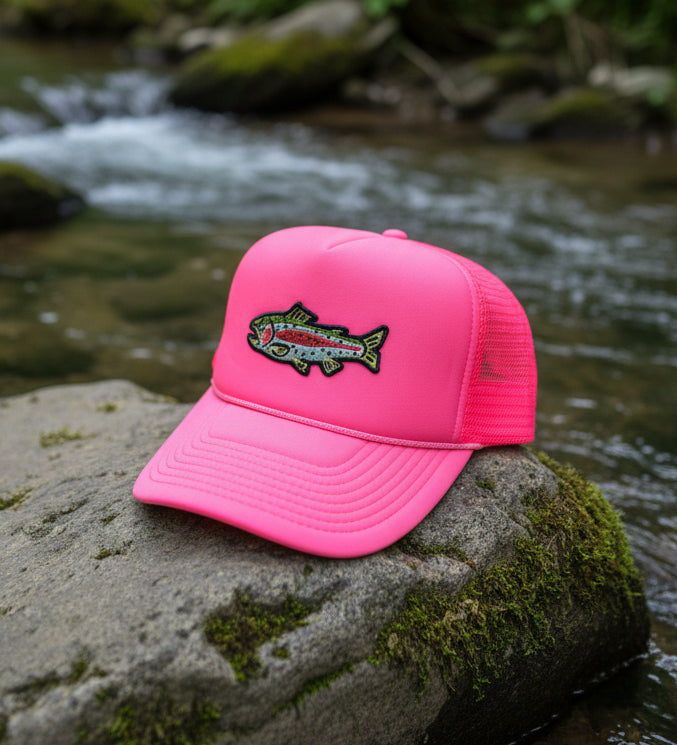 A bright pink trucker cap with a colorful embroidered trout patch on the front, resting on a gray textured surface.