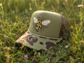 An olive green camouflage trucker hat with a detailed embroidered bee on the front panel, resting on a gray textured surface.