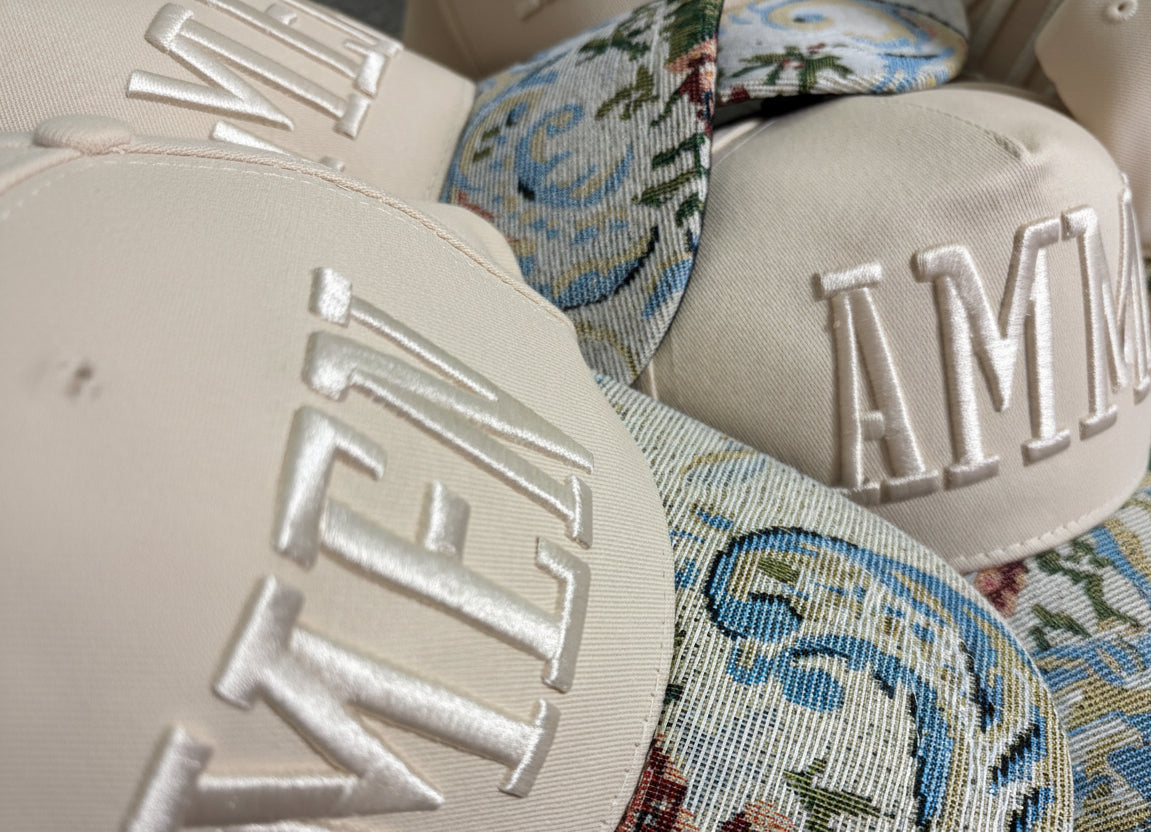 Close-up of beige baseball caps with large silver embroidered letters, layered over patterned fabric featuring blue, green, and red floral motifs.