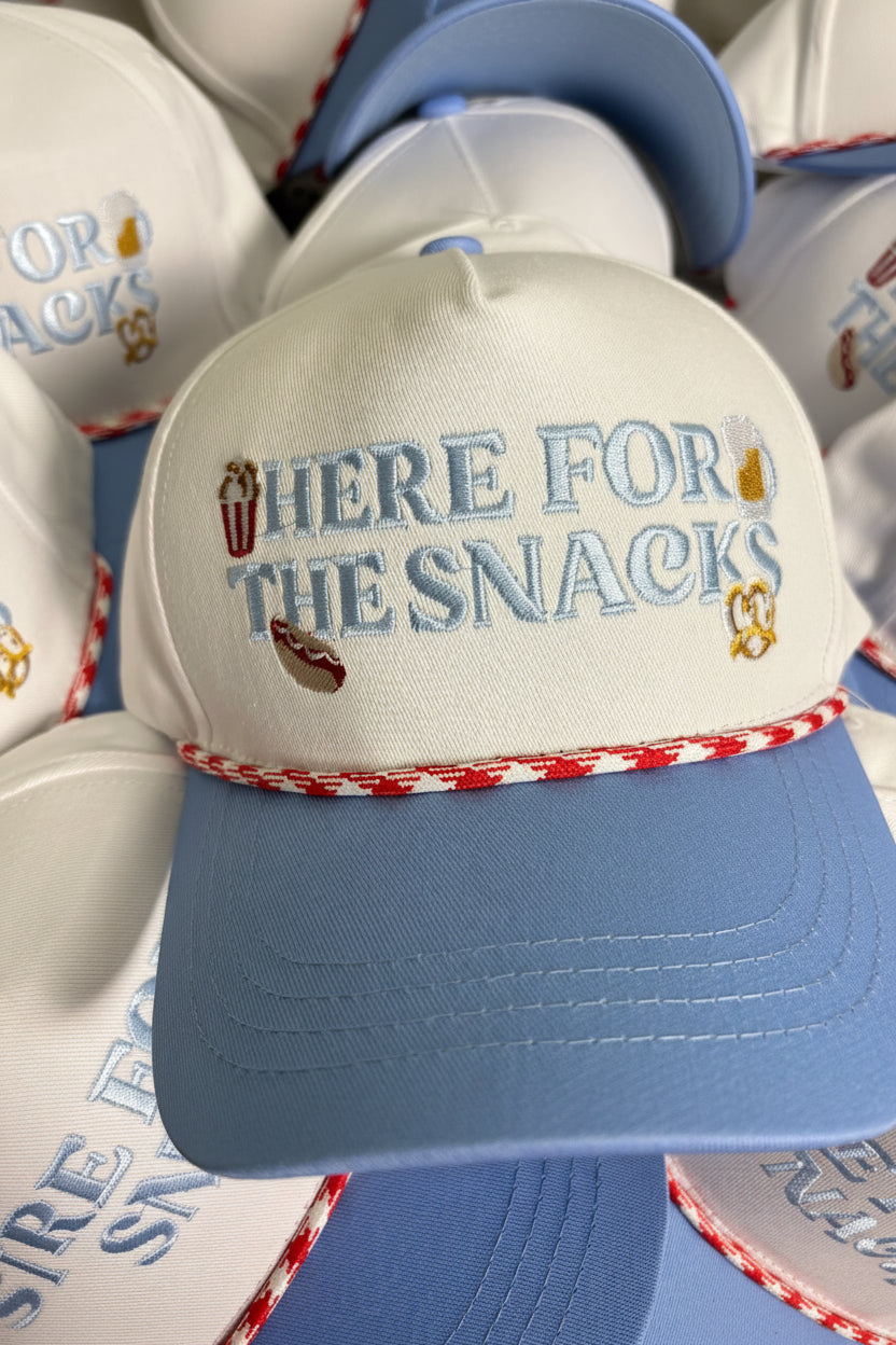 A white and light blue baseball cap with 'HERE FOR THE SNACKS' embroidered on the front, surrounded by other matching caps.