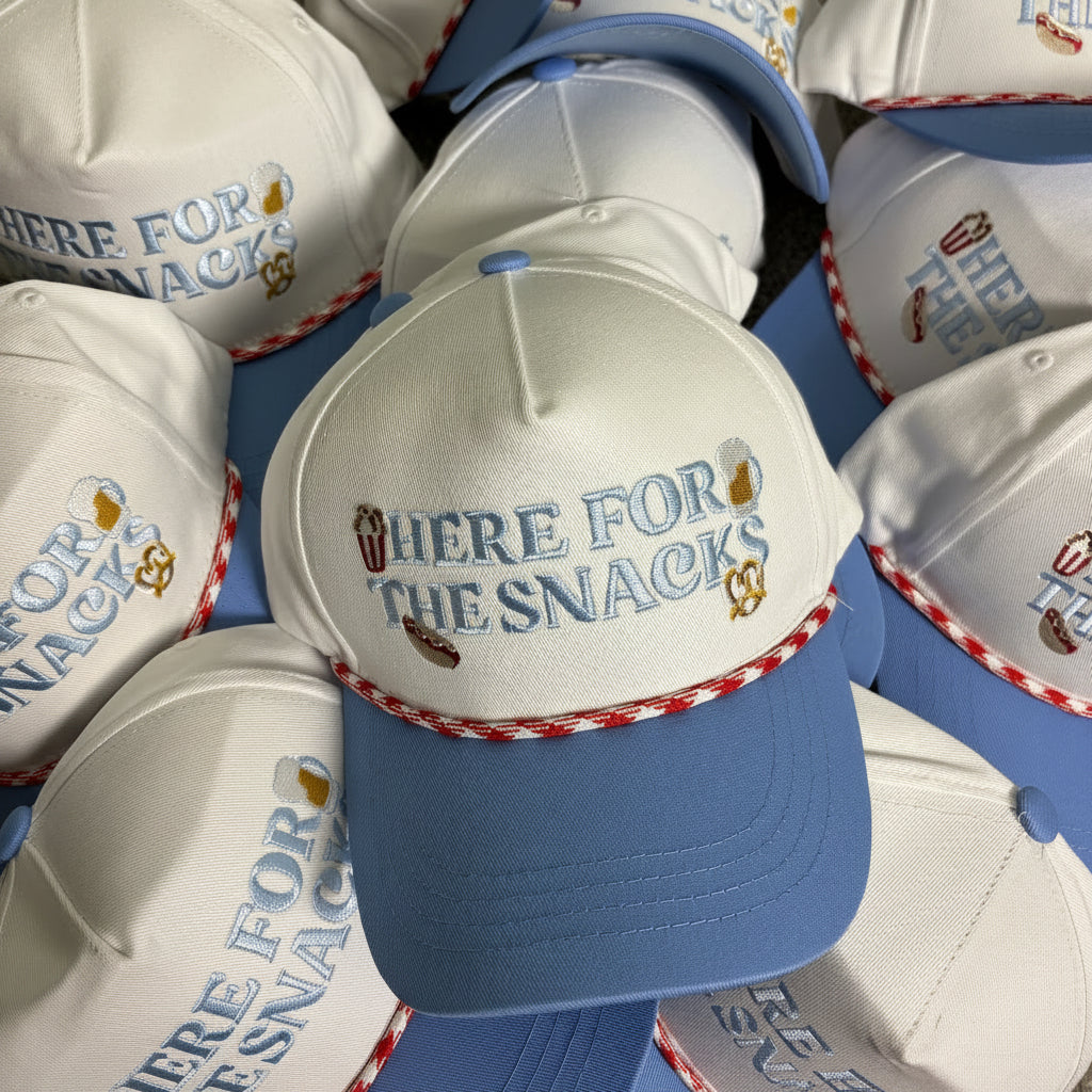 A pile of white and blue snapback hats with 'HERE FOR THE SNACKS' embroidered on the front, arranged on a beige carpet.