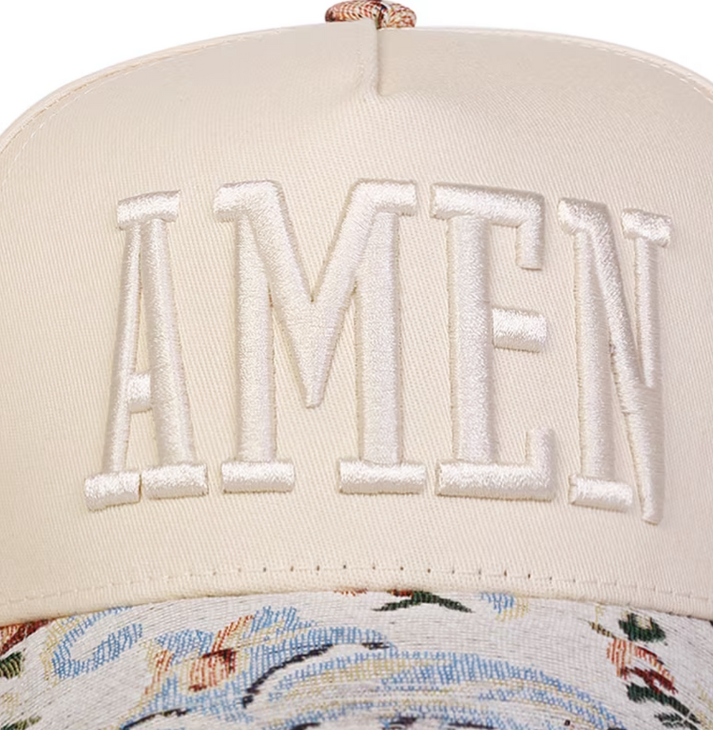A close-up of a cream-colored baseball cap featuring large white embroidered letters spelling 'AMEN' on the front, with a patterned fabric band visible below.
