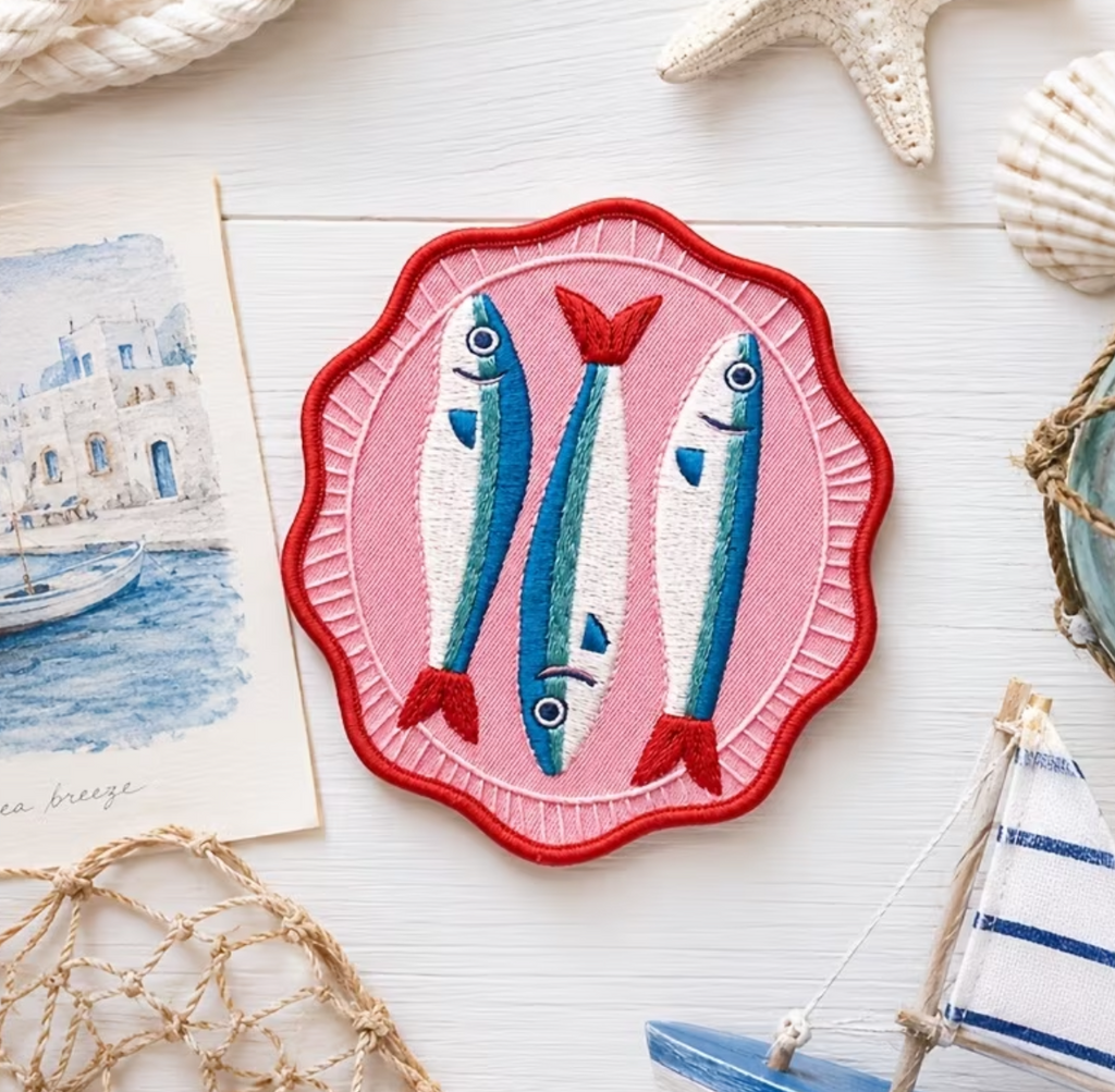 A square embroidered patch featuring two blue fish with red tails on a pink scalloped background, styled on a white surface with nautical props including a starfish, seashells, a net, and a small boat.
