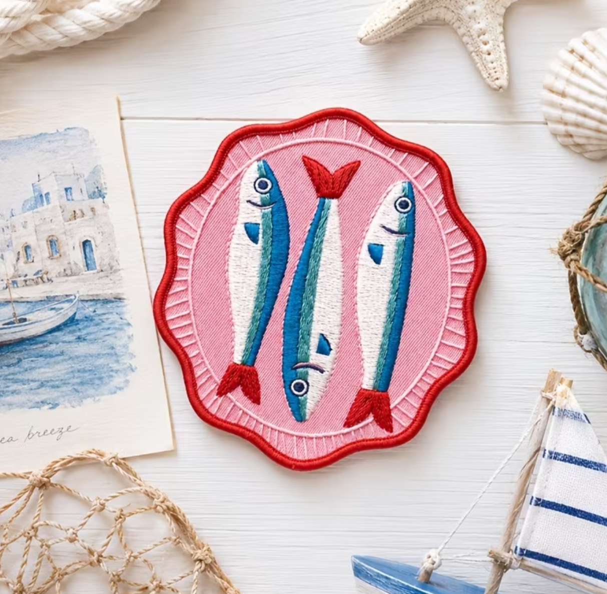 A square embroidered patch featuring two blue fish with red tails on a pink scalloped background, styled on a white surface with nautical props including a starfish, seashells, a net, and a small boat.