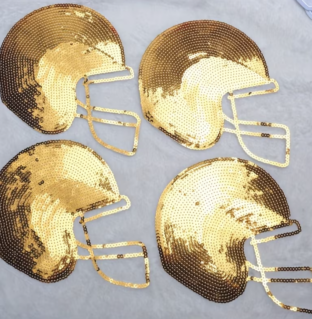 Four gold sequined football helmet decorations arranged in a 2x2 grid on a gray surface, shown within a phone screenshot with black bars and app interface elements.