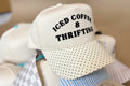 A cream-colored baseball cap with black embroidered text reading 'ICED COFFEE & THRIFTING' on the front panel, featuring a polka-dotted visor. The cap is shown resting on other hats in a pile.