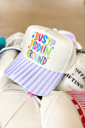 A cream-colored trucker-style cap with a pastel lavender and white striped brim, featuring colorful embroidered text that reads 'BUSHY PADDLIN' AROUND' on the front panel.