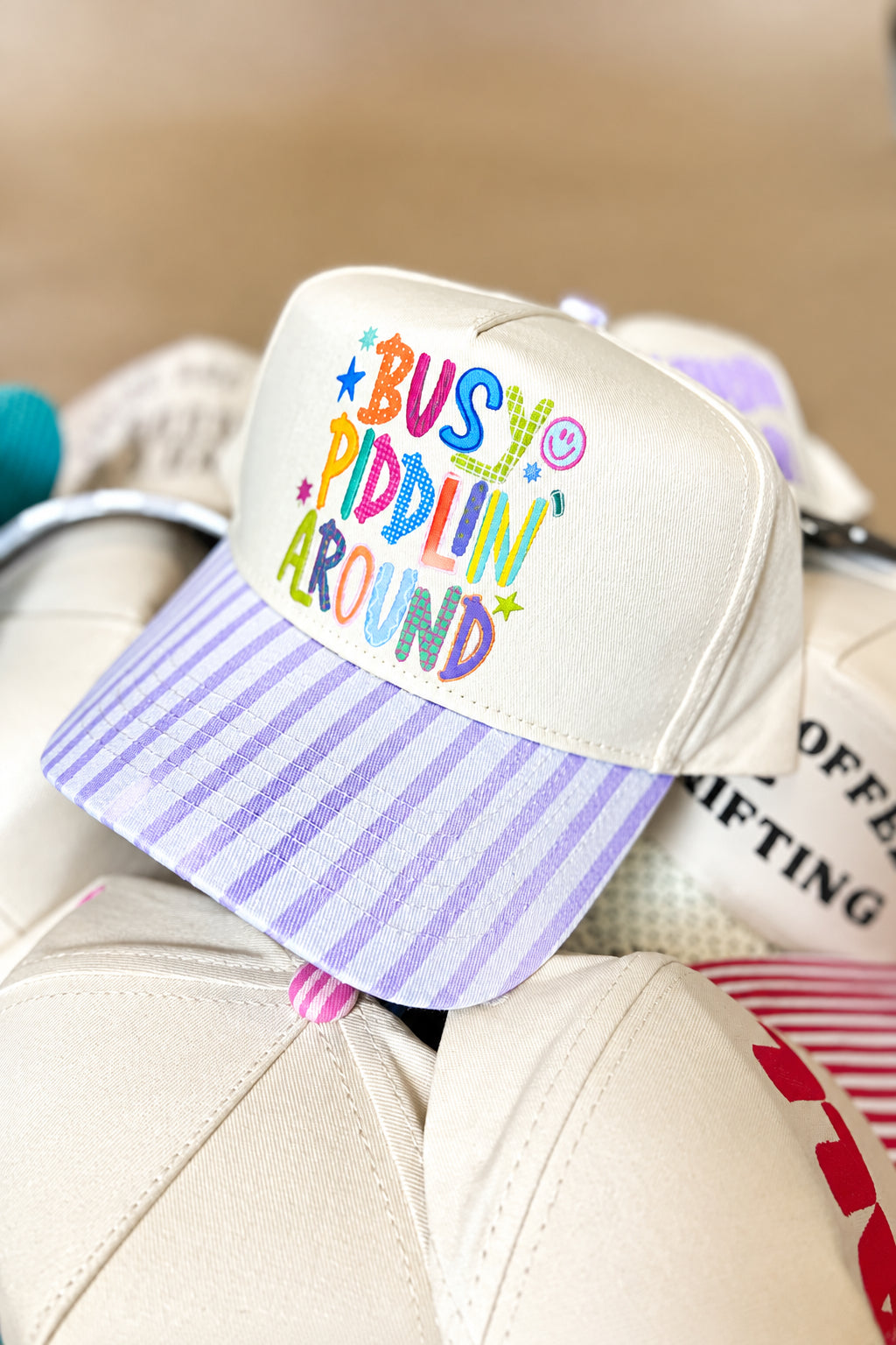 A cream-colored trucker-style cap with a pastel lavender and white striped brim, featuring colorful embroidered text that reads 'BUSHY PADDLIN' AROUND' on the front panel.