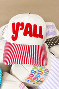 A cream-colored snapback cap with red embroidered 'y'ALL' text on the front and a red-and-white striped visor, shown resting on top of other hats.