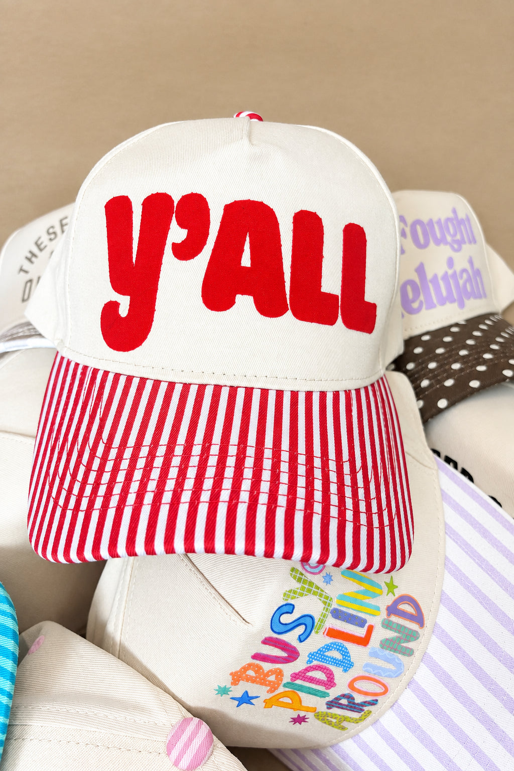 A cream-colored snapback cap with red embroidered 'y'ALL' text on the front and a red-and-white striped visor, shown resting on top of other hats.