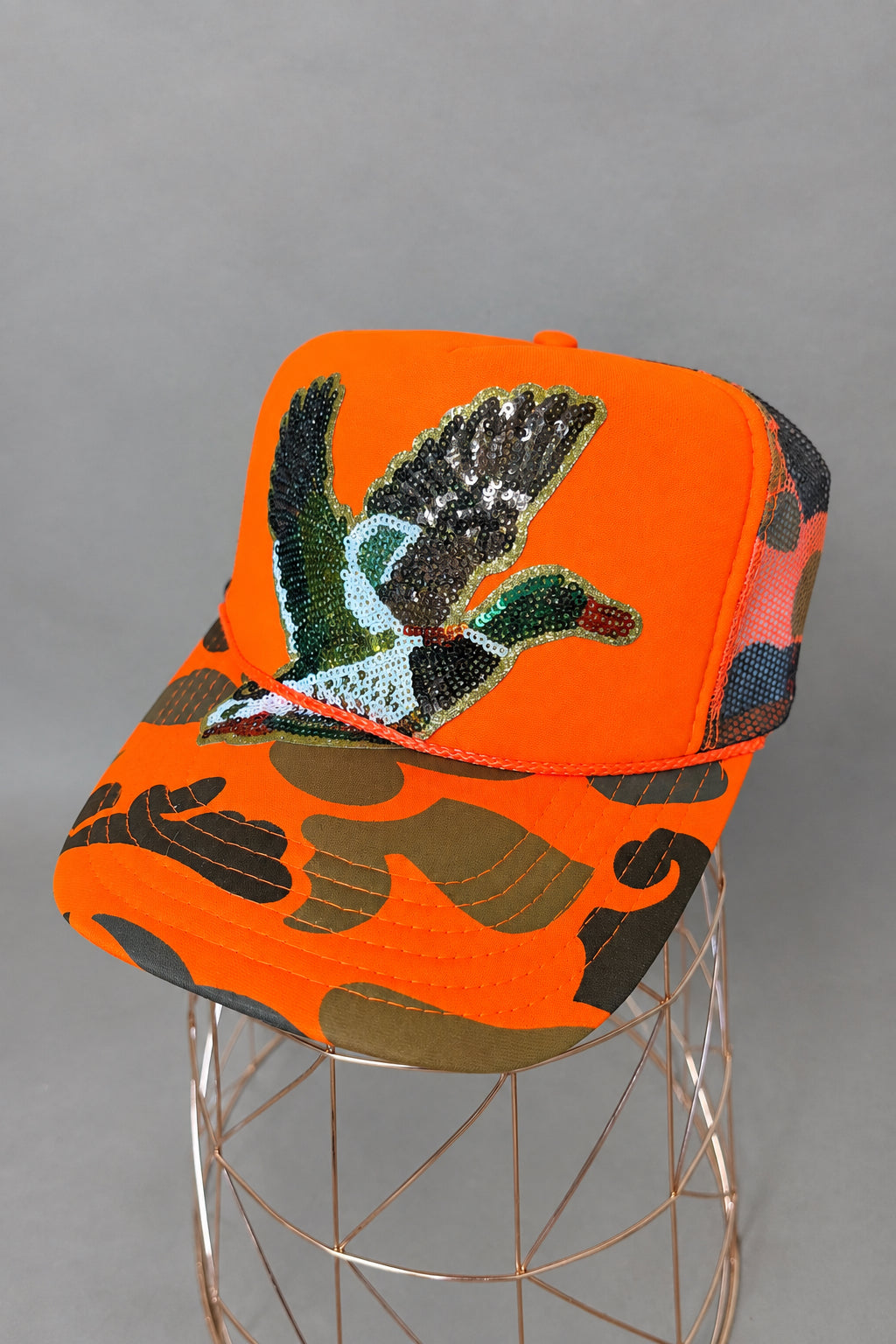 Bright orange trucker cap with a sequined duck graphic on the front and a neon orange camouflage brim, displayed on a wire stand against a neutral gray background.